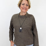 A woman with short blonde hair wearing a textured, ribbed brown long-sleeve sweater and light blue jeans, standing against a plain white background.