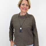A woman with short blonde hair wearing a textured, ribbed brown long-sleeve sweater and light blue jeans, standing against a plain white background.