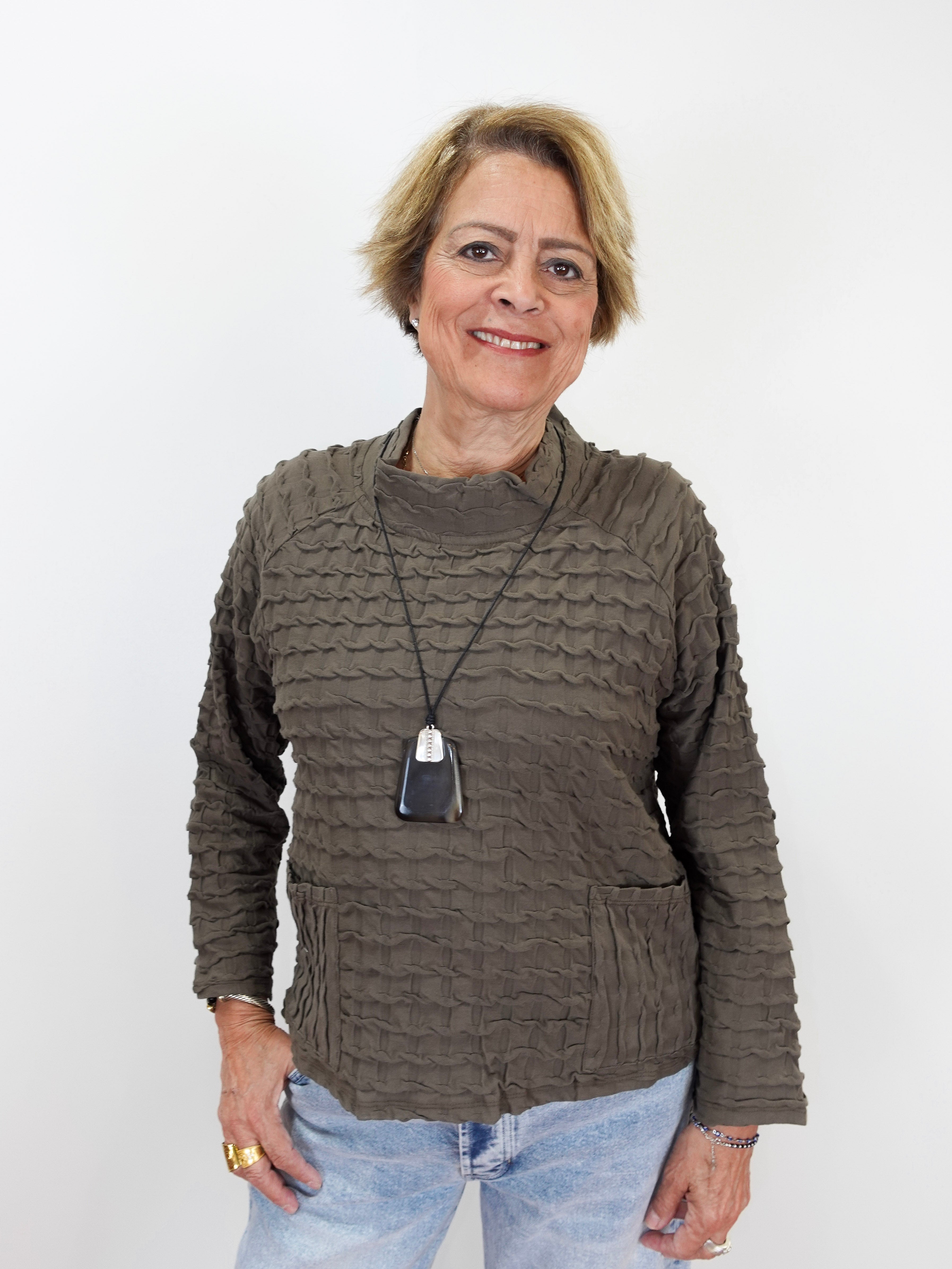 A woman with short blonde hair wearing a textured, ribbed brown long-sleeve sweater and light blue jeans, standing against a plain white background.