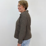 A woman with short blonde hair is shown in profile wearing a textured, ribbed knit cropped jacket in a dark brownish-gray color, paired with light blue jeans, against a plain light gray background.