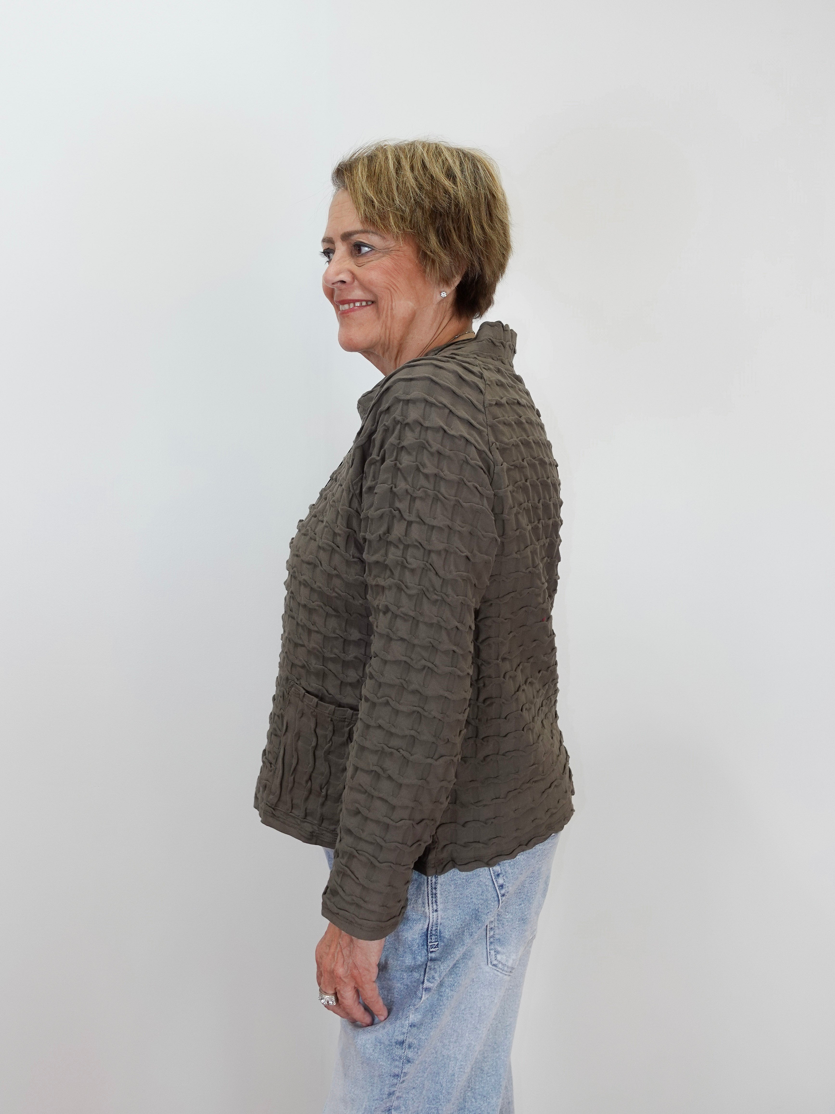 A woman with short blonde hair is shown in profile wearing a textured, ribbed knit cropped jacket in a dark brownish-gray color, paired with light blue jeans, against a plain light gray background.