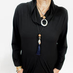 A woman wearing a black long-sleeve cowl-neck dress or tunic top, styled with a long beaded necklace featuring a silver ring, wooden beads, and a blue tassel.