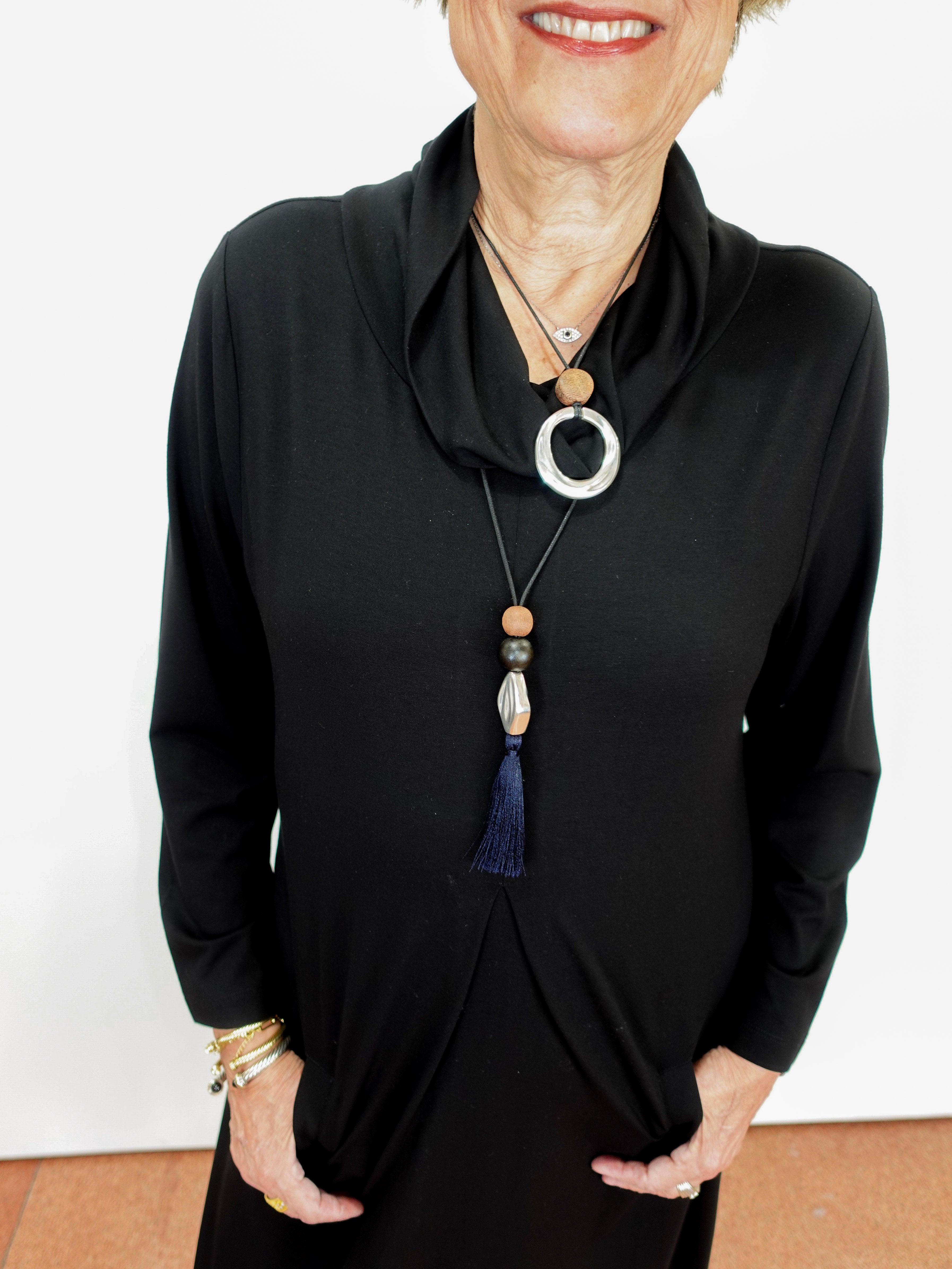 A woman wearing a black long-sleeve cowl-neck dress or tunic top, styled with a long beaded necklace featuring a silver ring, wooden beads, and a blue tassel.