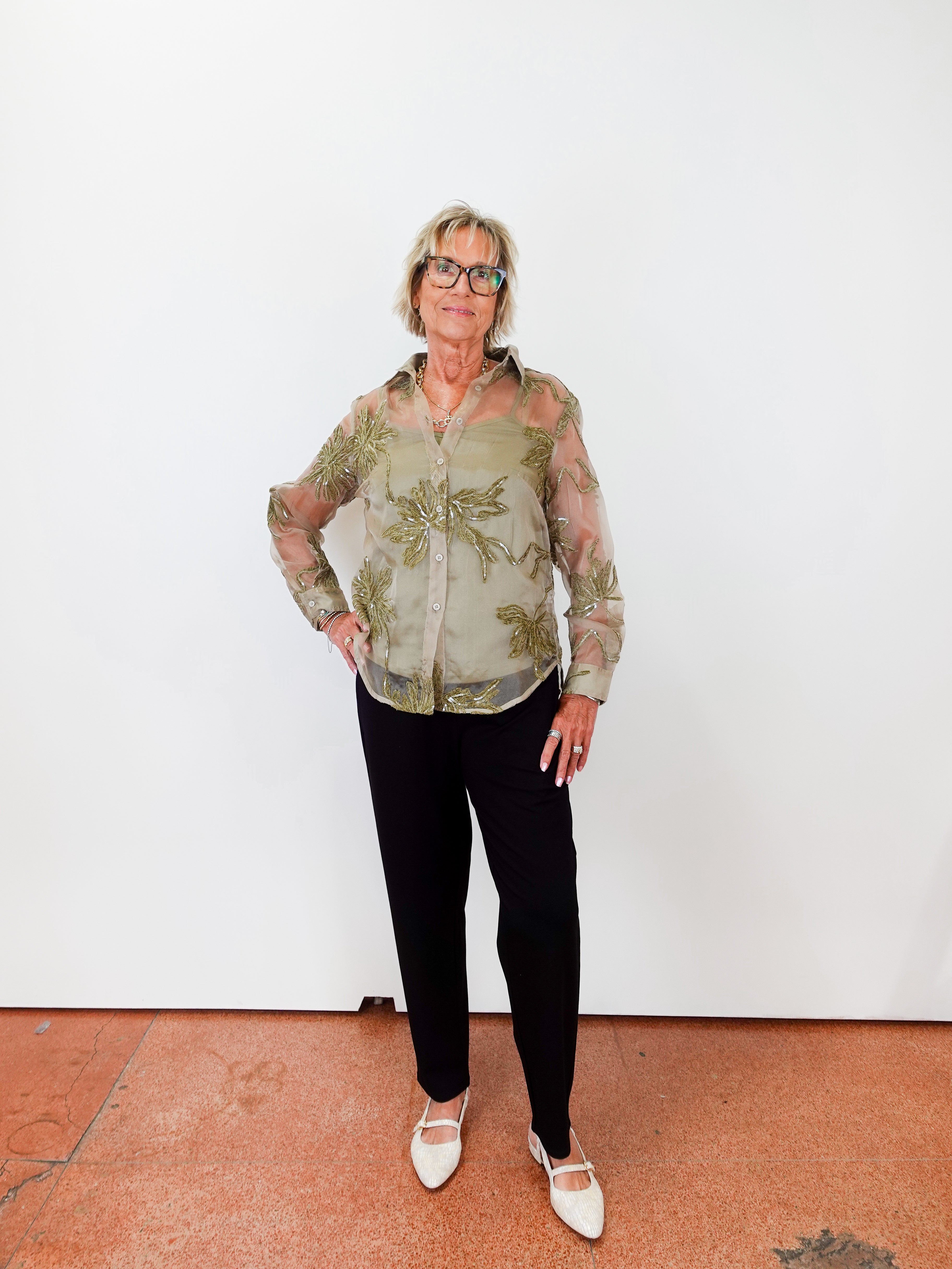 Person wearing a patterned shirt and black pants standing against a white wall.