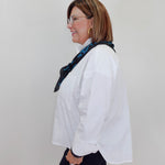 A woman in profile wearing a white long-sleeve button-up shirt, a dark navy patterned scarf, and dark pants, standing against a plain light gray wall.