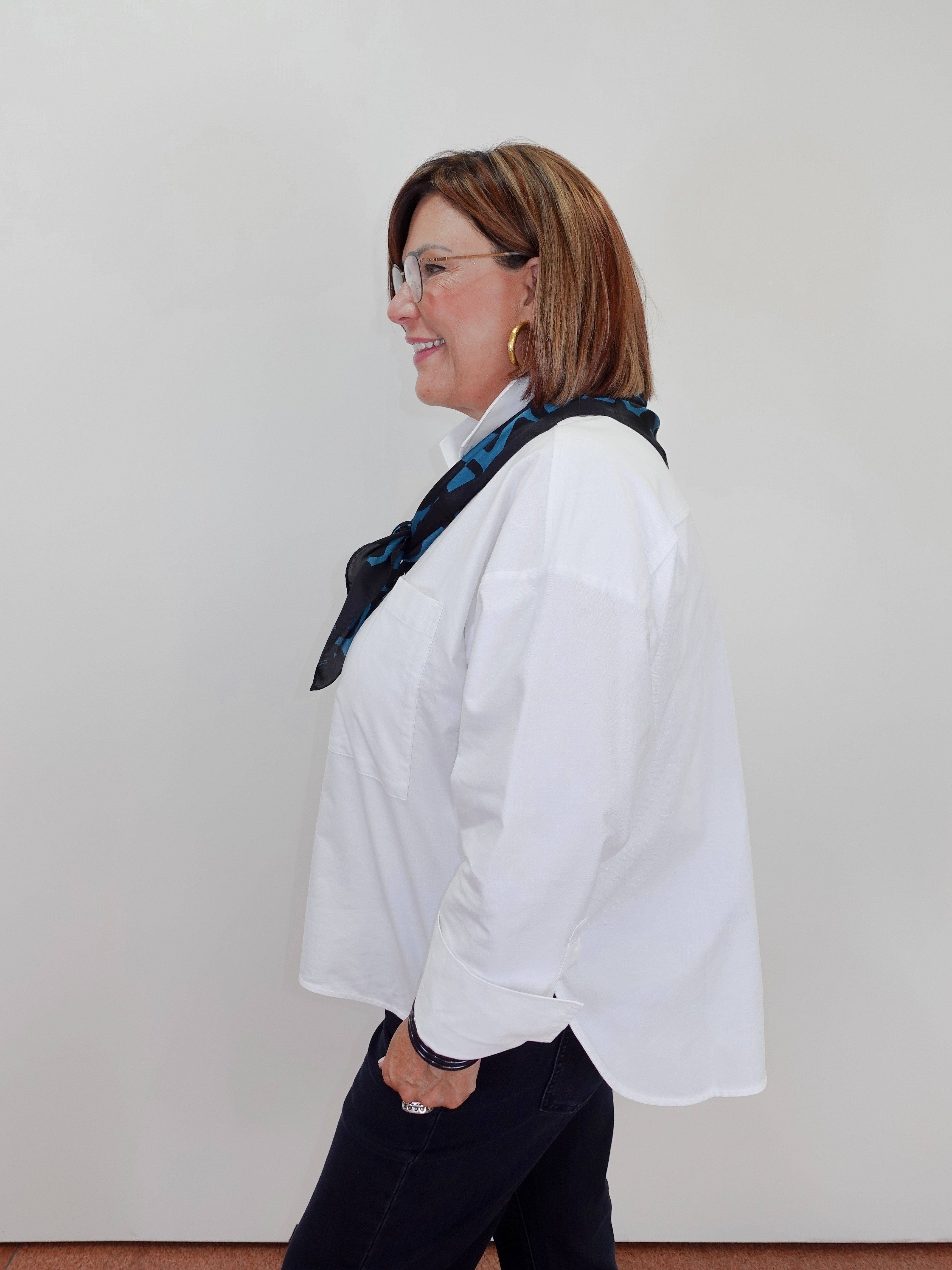 A woman in profile wearing a white long-sleeve button-up shirt, a dark navy patterned scarf, and dark pants, standing against a plain light gray wall.