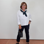 A woman modeling a white long-sleeve button-up shirt with a dark patterned neck scarf, black pants, and dark gray peep-toe flats, standing against a plain white wall.