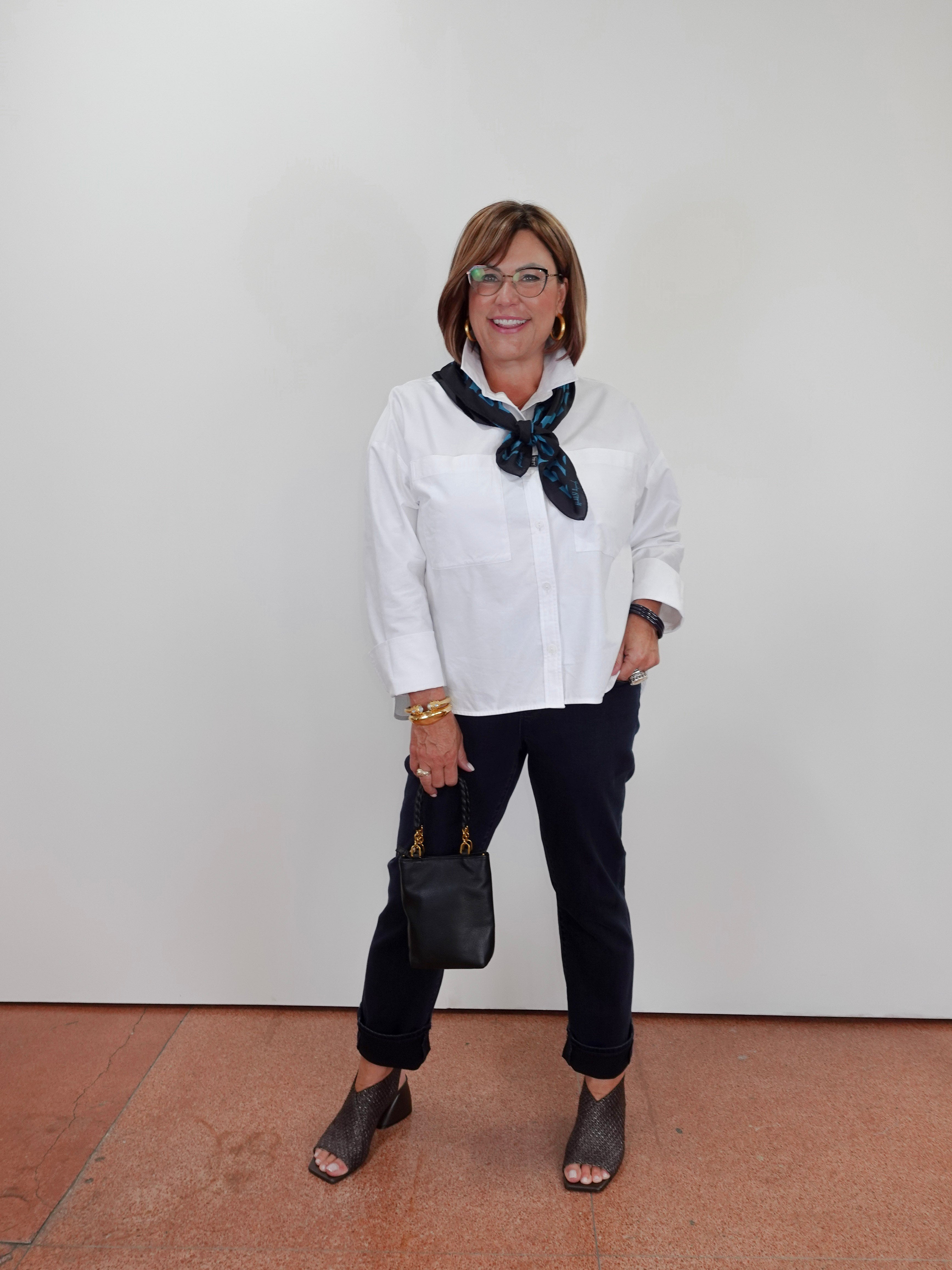 A woman modeling a white long-sleeve button-up shirt with a dark patterned neck scarf, black pants, and dark gray peep-toe flats, standing against a plain white wall.