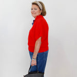 Woman in a red sweater and blue jeans holding a gray handbag against a white wall.
