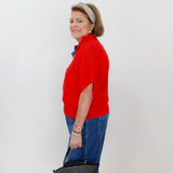 Woman in a red sweater and blue jeans holding a gray handbag against a white wall.