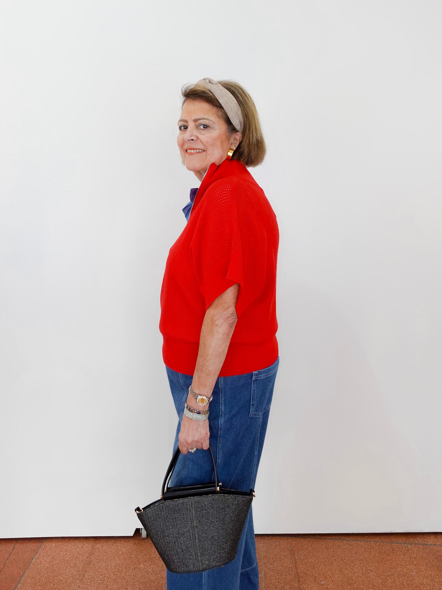 Woman in a red sweater and blue jeans holding a gray handbag against a white wall.