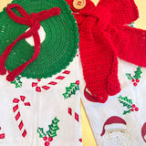 Crocheted Christmas decorations including a green wreath and red bow on a fabric background with holiday patterns.
