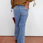 A person shown from the back wearing light-wash blue flare jeans with decorative side embellishments, a patterned olive-brown blouse, and black heels, standing against a plain white wall.