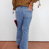 A person shown from the back wearing light-wash blue flare jeans with decorative side embellishments, a patterned olive-brown blouse, and black heels, standing against a plain white wall.
