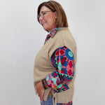 A woman wearing a beige knit sweater vest over a colorful patterned blouse and blue jeans, standing against a plain light gray background.
