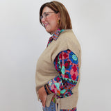 A woman wearing a beige knit sweater vest over a colorful patterned blouse and blue jeans, standing against a plain light gray background.