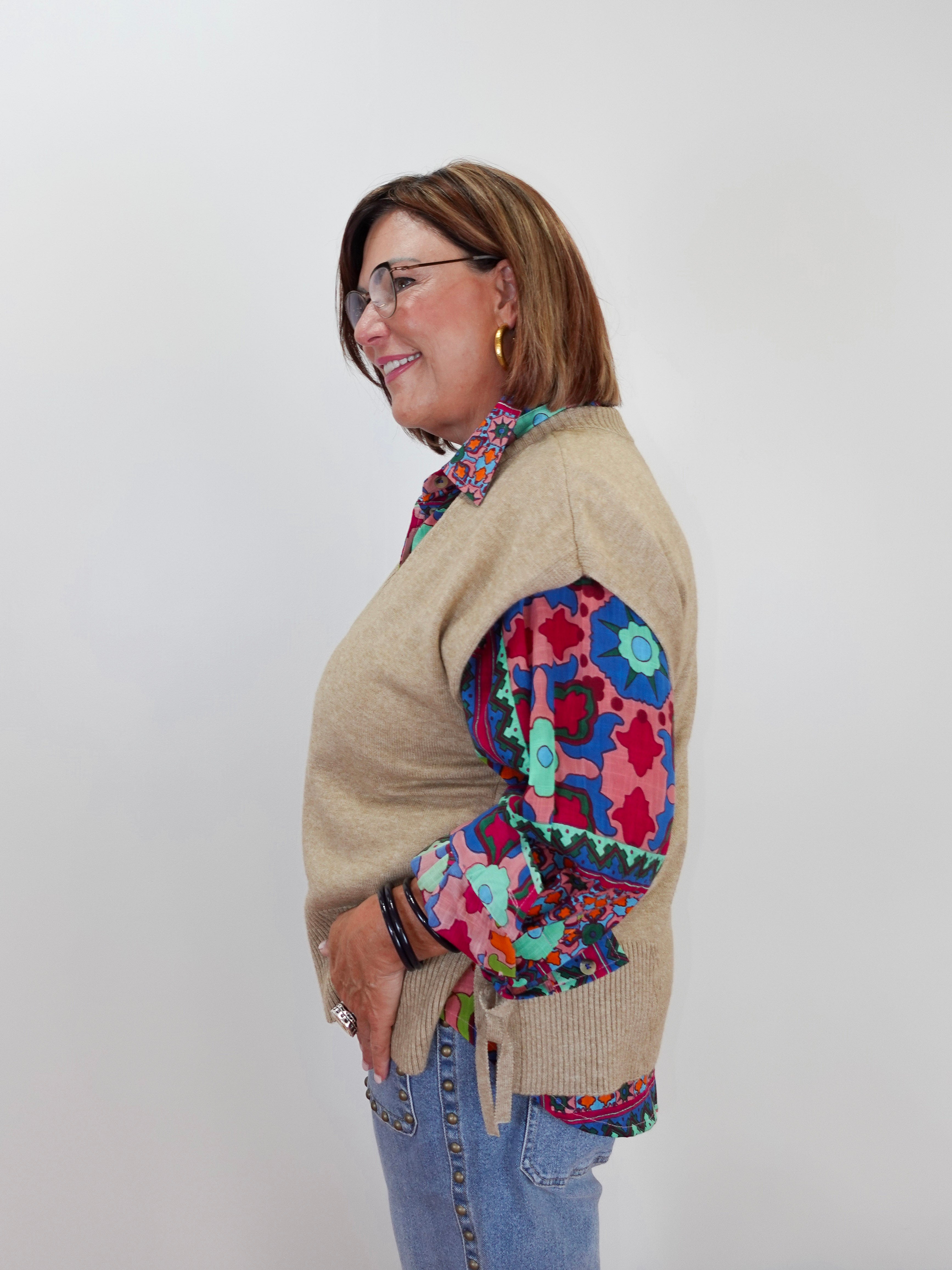 A woman wearing a beige knit sweater vest over a colorful patterned blouse and blue jeans, standing against a plain light gray background.