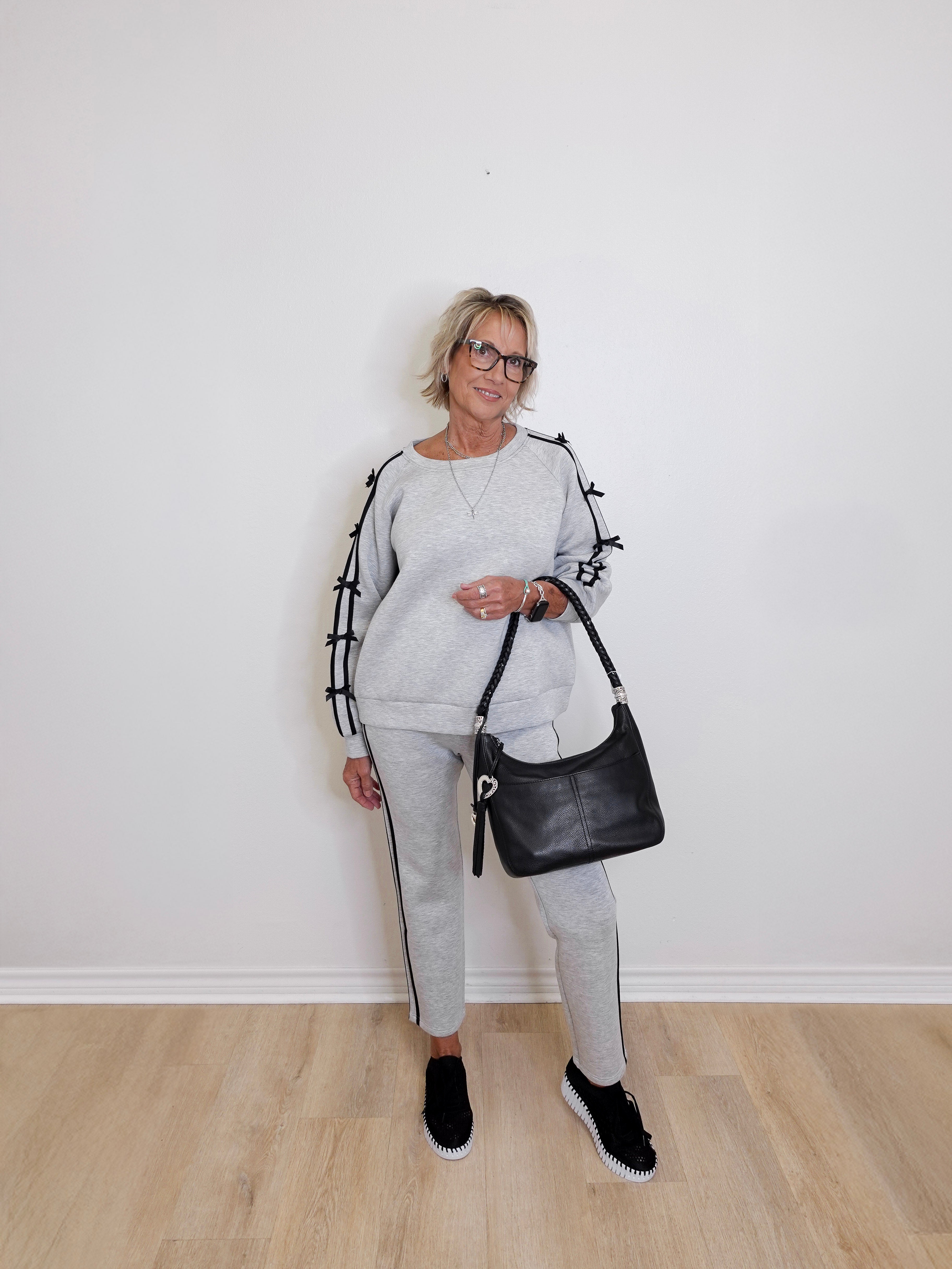 Woman in gray tracksuit holding a black handbag against a white wall