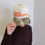 Person wearing a cap with 'HE'S HUNTING' text against a plain background