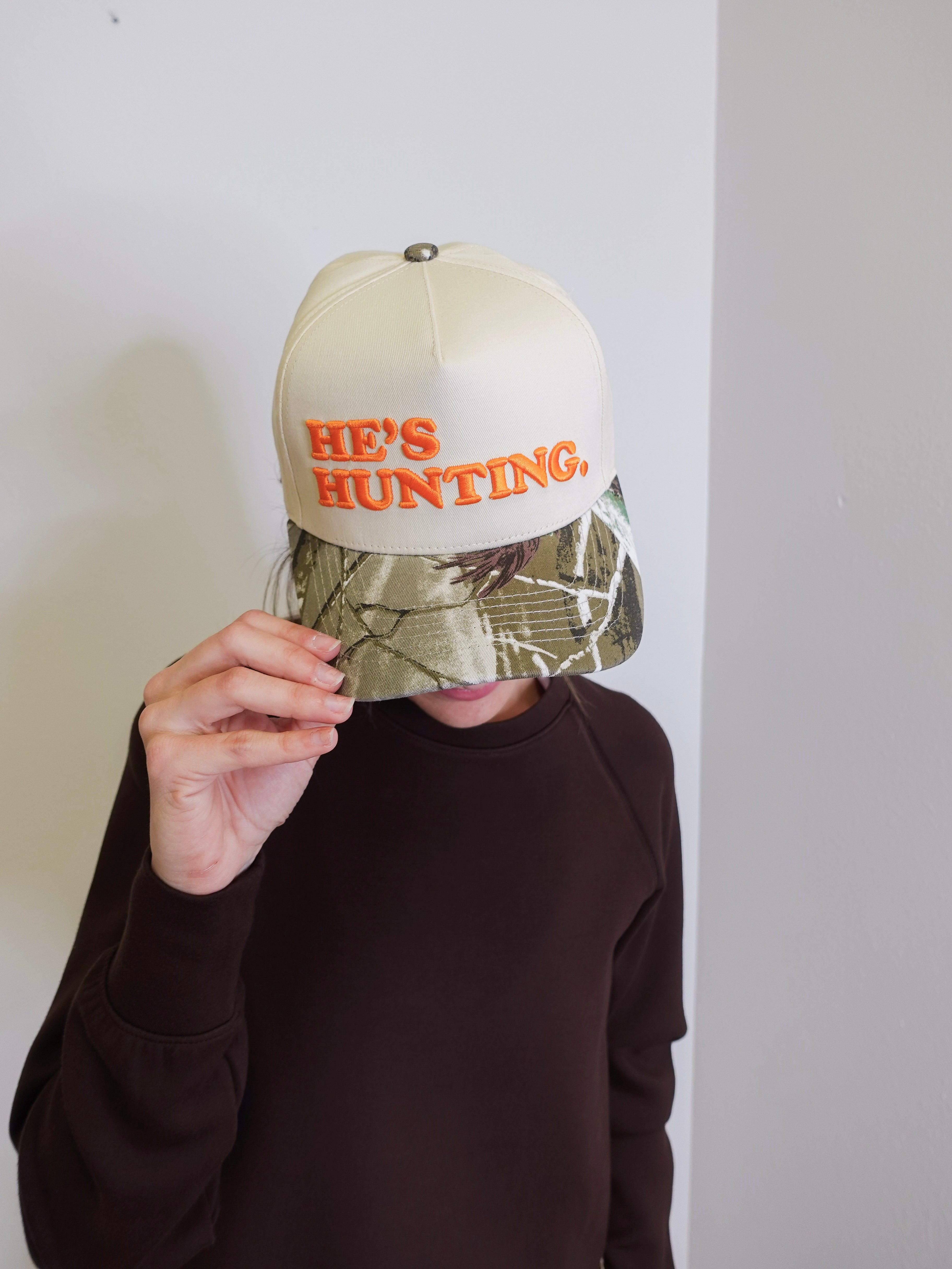 Person wearing a cap with 'HE'S HUNTING' text against a plain background