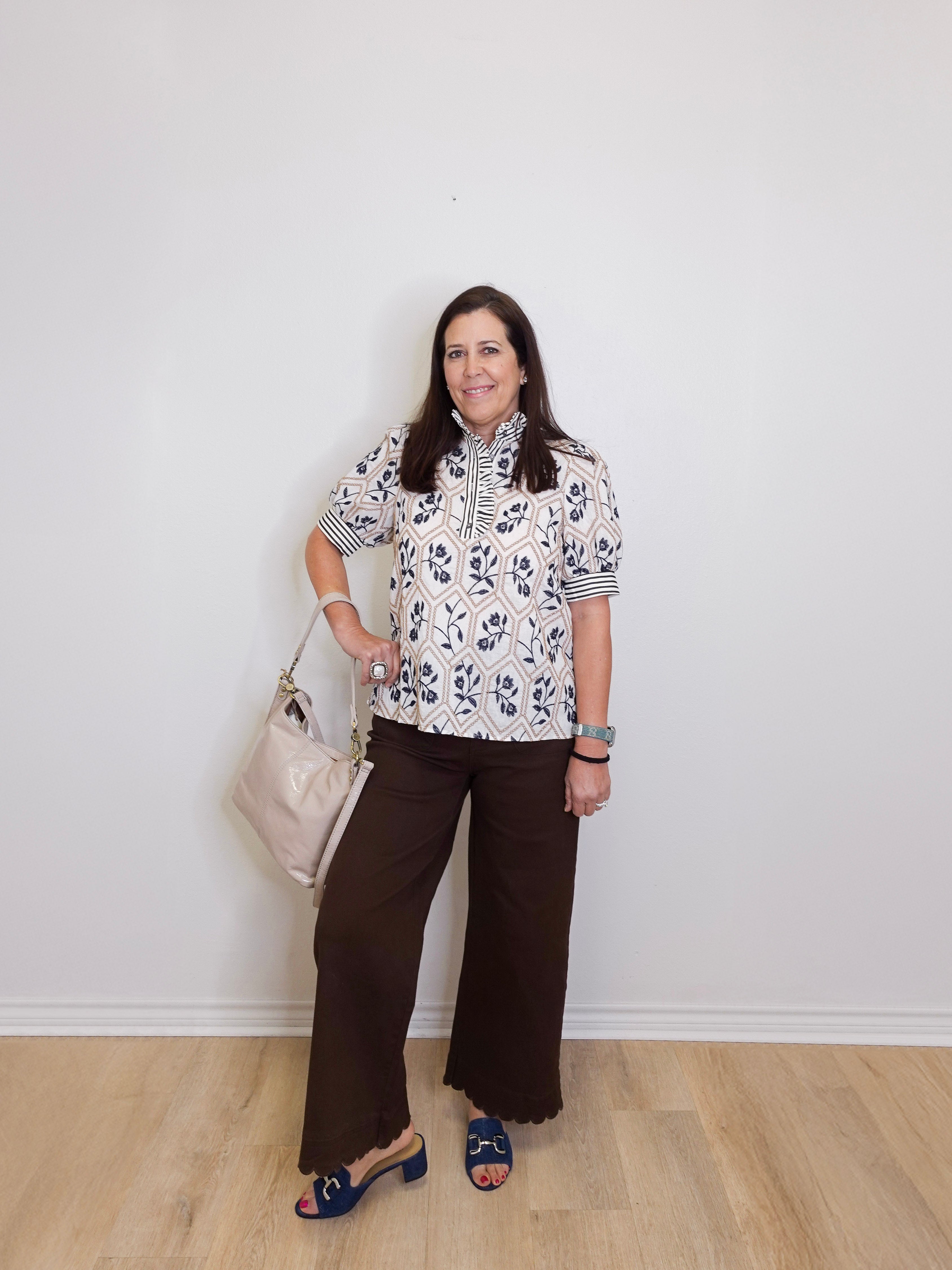 Woman wearing a floral blouse and brown pants against a white wall.