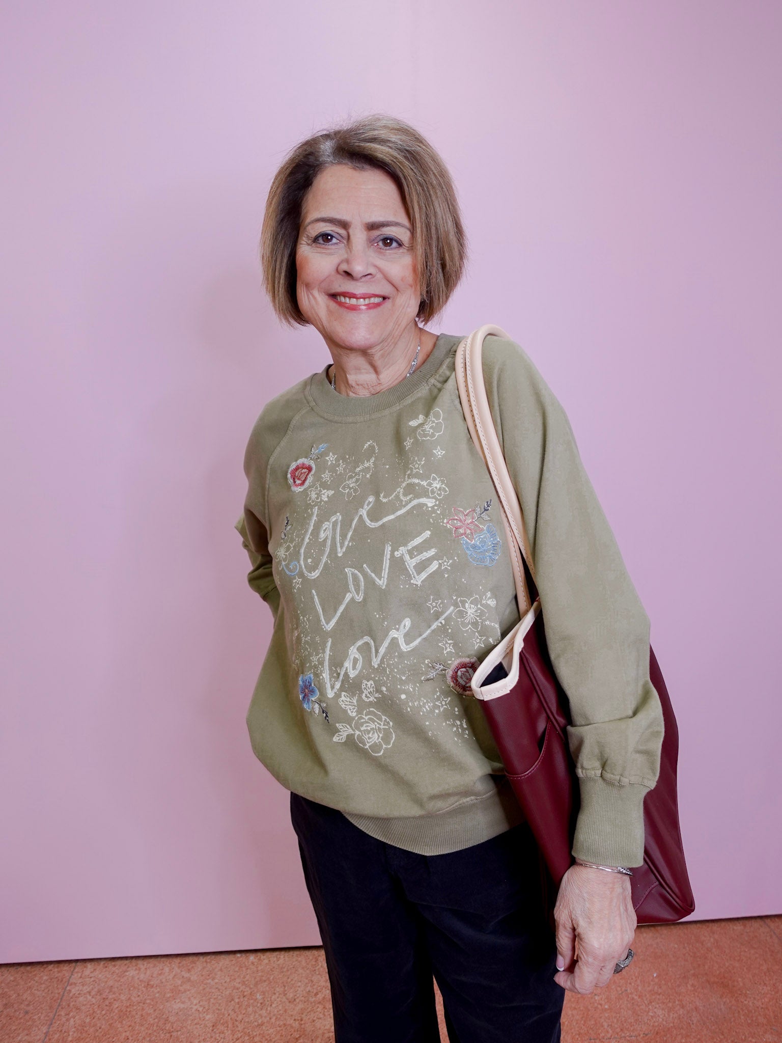Woman wearing a green sweatshirt with text and graphics, holding a red bag against a pink background