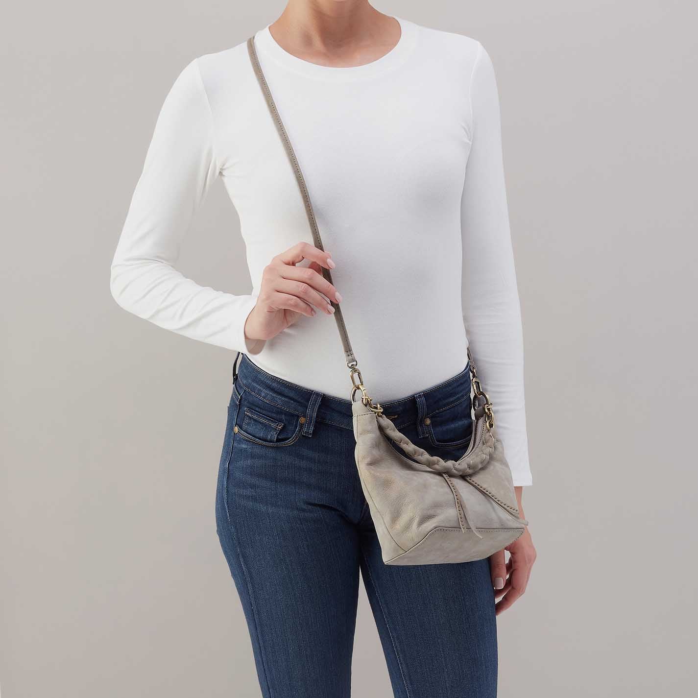 Person wearing a white long-sleeve shirt and blue jeans, holding a beige crossbody bag against a gray background