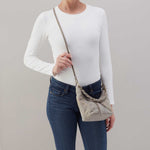 Person wearing a white long-sleeve shirt and blue jeans, holding a beige crossbody bag against a gray background