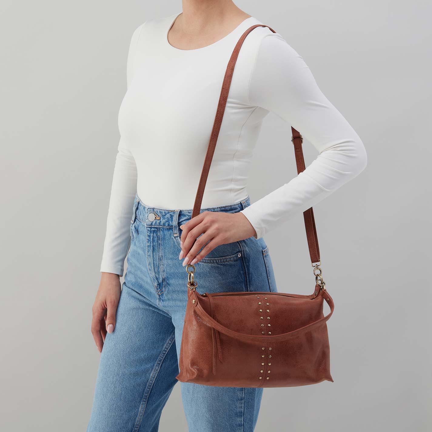 Person wearing a white long-sleeve top and blue jeans, holding a brown leather handbag against a gray background