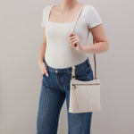 Person wearing a white top and blue jeans holding a beige crossbody bag against a plain background