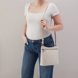 Person wearing a white top and blue jeans holding a beige crossbody bag against a plain background