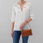 Person wearing a white blouse with floral patterns and blue jeans, holding a brown leather handbag against a gray background.