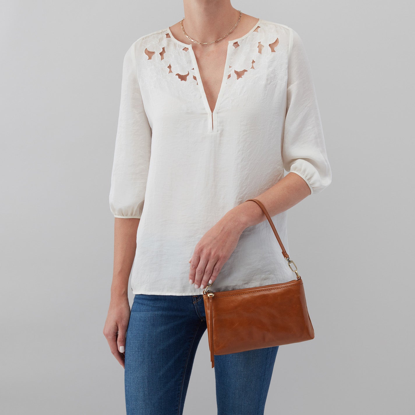 Person wearing a white blouse with floral patterns and blue jeans, holding a brown leather handbag against a gray background.