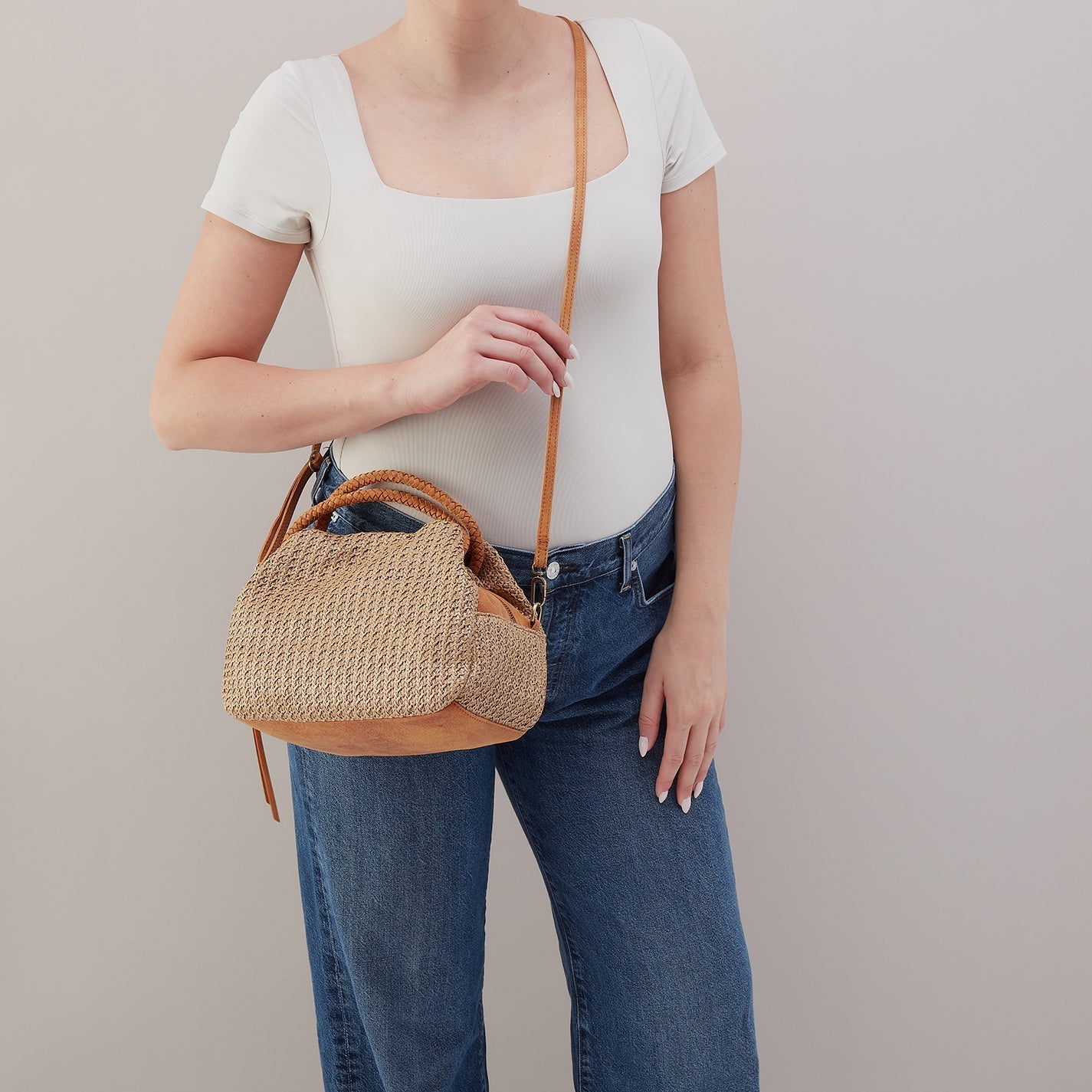 Person wearing a white top and blue jeans, holding a woven handbag against a plain background