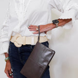 Person wearing a white shirt, blue jeans, and holding a brown leather bag on a plain background