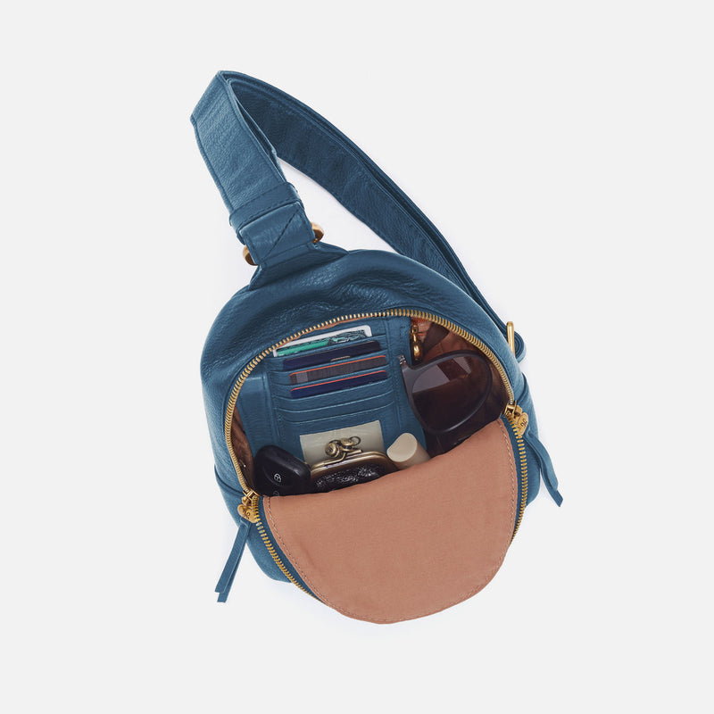 Blue sling bag with a brown interior, containing items like a phone and wallet, on a white background.