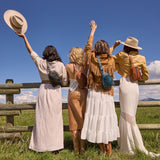 Four women in vintage-style clothing standing in a field with a wooden fence and blue sky.