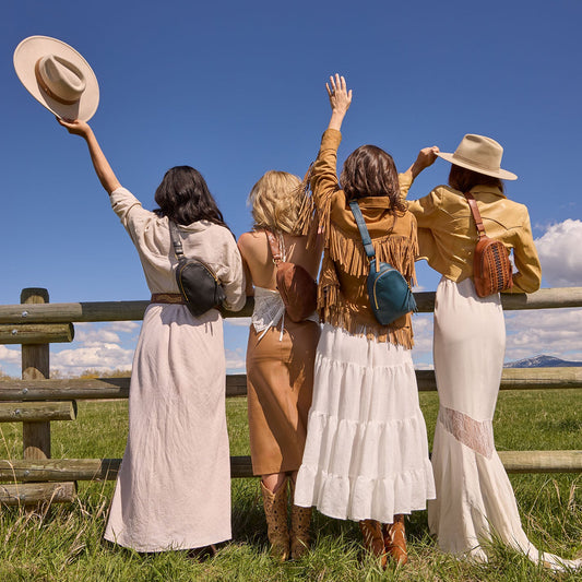 Four women in vintage-style clothing standing in a field with a wooden fence and blue sky.