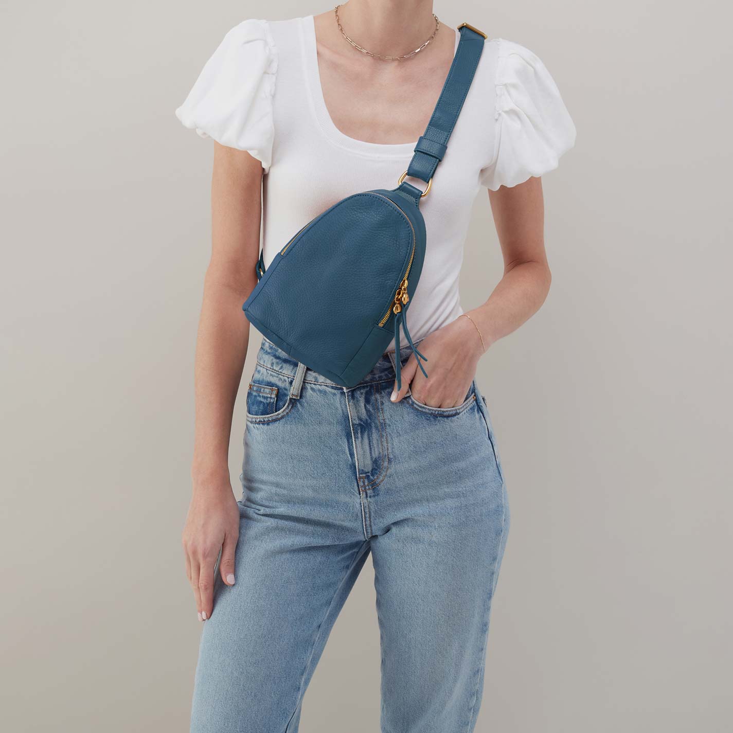 Person wearing a white top and blue jeans with a teal crossbody bag on a plain background