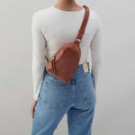 Person wearing a brown leather crossbody bag with a plain background