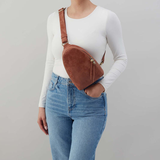 Person wearing a white long-sleeve shirt, blue jeans, and a brown suede crossbody bag on a gray background