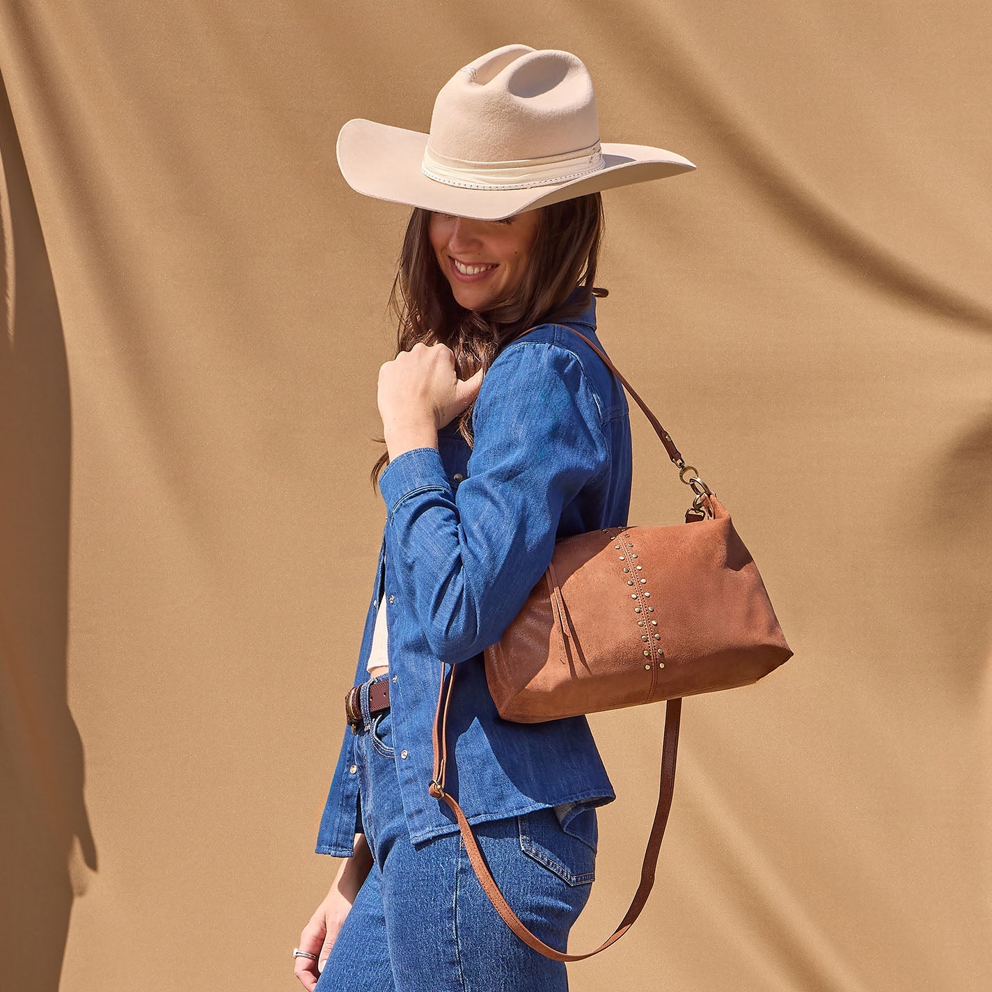 Woman wearing a blue denim outfit with a brown leather handbag against a beige background