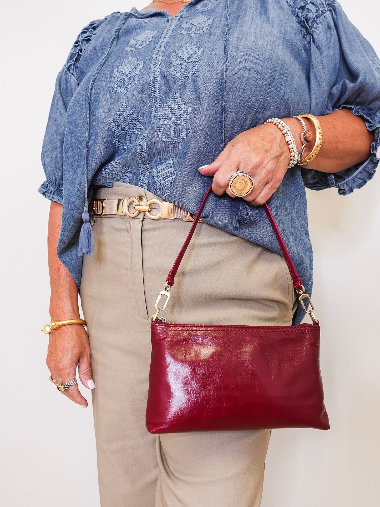 Person wearing a blue embroidered blouse, beige pants, and gold jewelry, holding a red handbag.