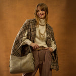 Woman in a plaid coat and brown pants holding a beige handbag against a brown background