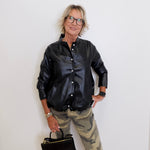 Person wearing a black leather jacket and camouflage pants holding a black handbag against a white wall.