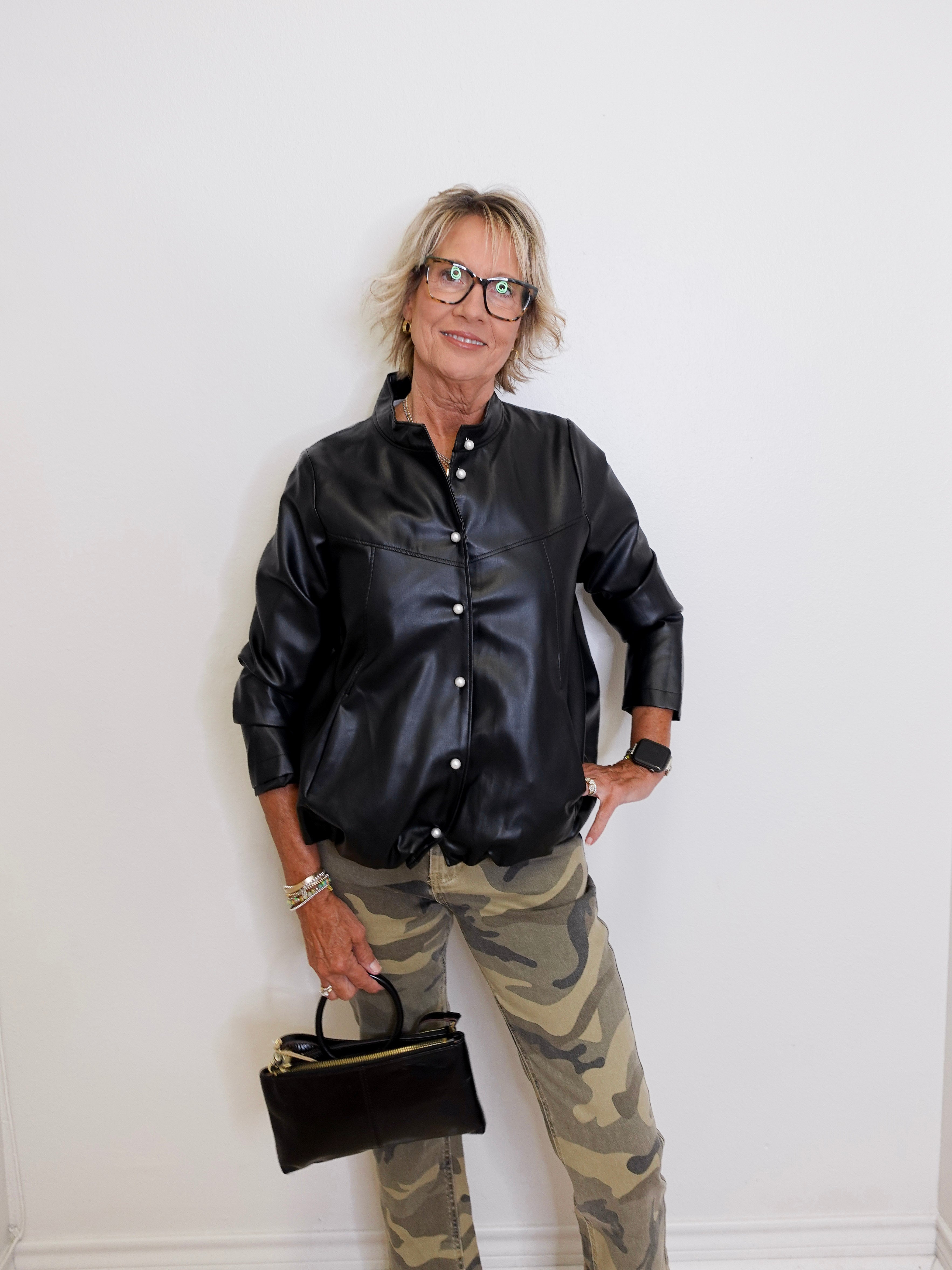 Person wearing a black leather jacket and camouflage pants holding a black handbag against a white wall.