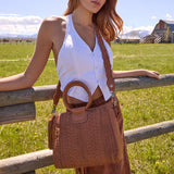 Woman holding a brown woven handbag in a field with a wooden fence.