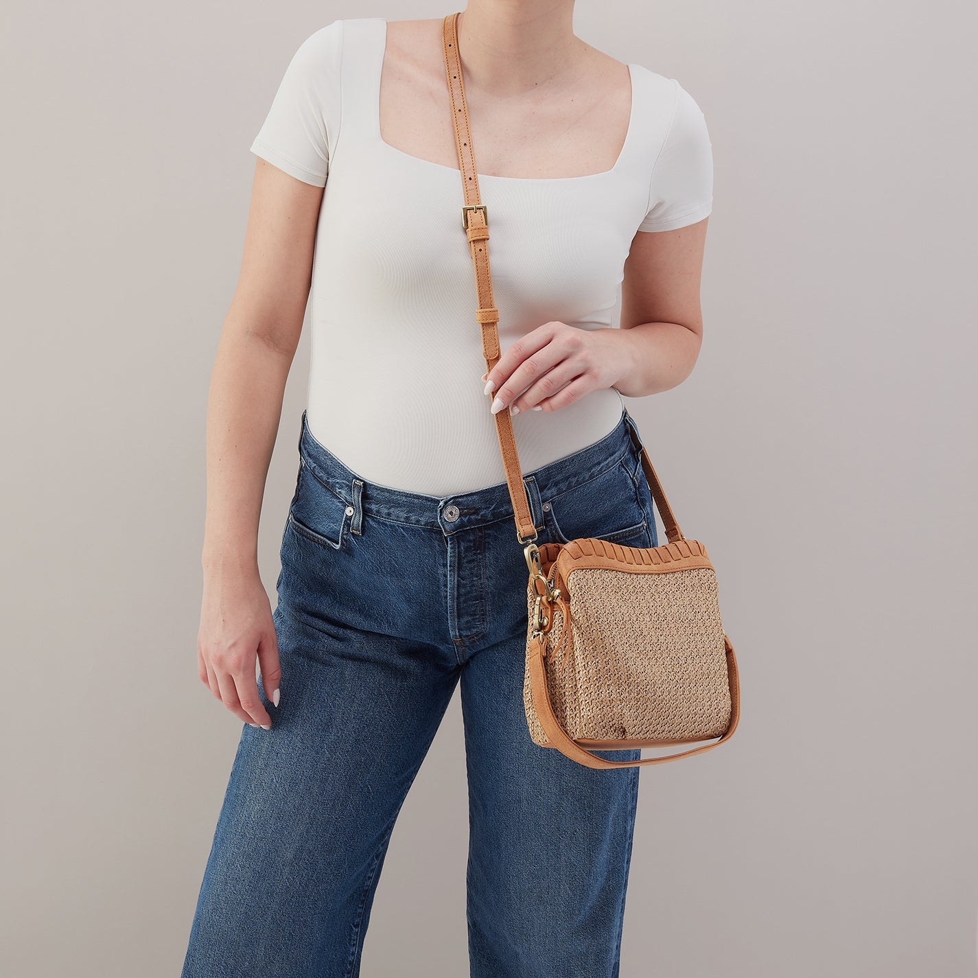 Person wearing a white top and blue jeans holding a woven handbag against a plain background