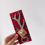 Decorative antler-shaped bottle opener on a red plaid card held by a hand against a white background