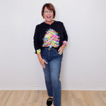 Person wearing a black sweater with a colorful design and blue jeans on a plain white background